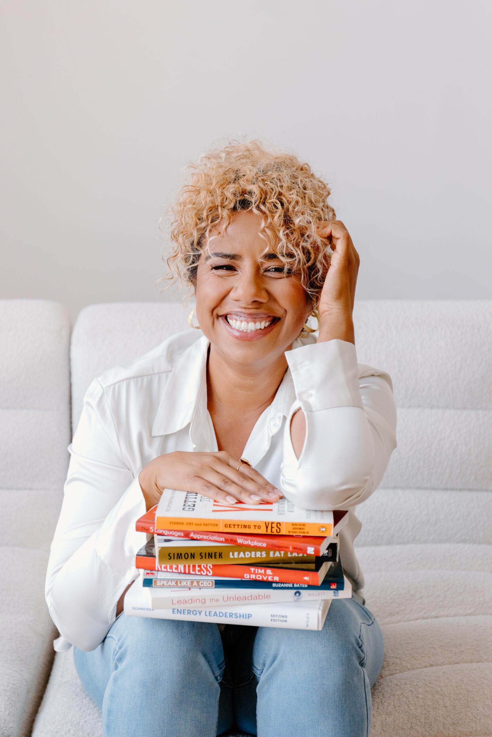 "Sharilyn Chavez, a Certified Professional Coach, smiling warmly while sitting on a couch with a stack of leadership and self-help books, including 'Getting to Yes' and 'Energy Leadership.'"